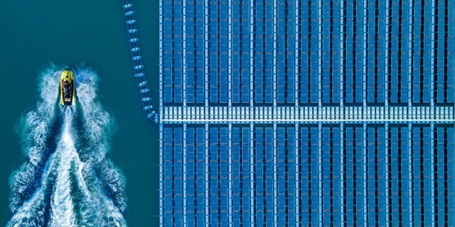 This aerial image captures a yellow boat moving through blue water, creating a wake as it passes beside a large array of floating solar panels. The solar panels are arranged in neat, geometric rows.