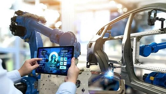 A person holds a tablet displaying data while overseeing a robotic arm assembling a car frame in a modern factory.