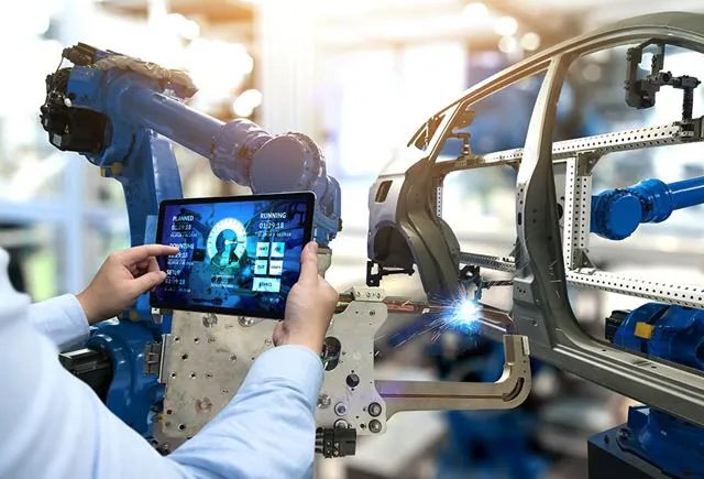 A person holds a tablet displaying data while overseeing a robotic arm assembling a car frame in a modern factory.