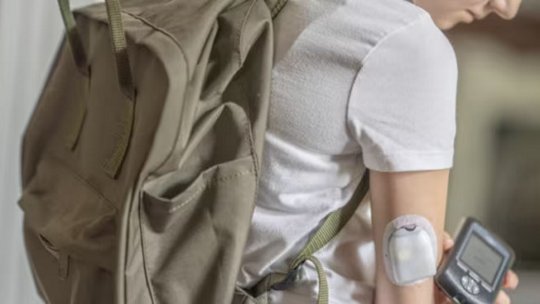A teenager wearing a white t-shirt and khaki backpack is shown from the side, displaying an insulin pump attached to their upper arm. The individual is holding a glucose monitoring device.