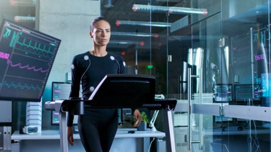 A person in a black sensor suit walks on a treadmill inside a modern laboratory or research facility. Multiple large monitors display heart rate and ECG data, with visible numbers such as 78 and 89:59.