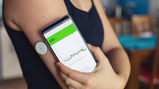 A person holds a smartphone displaying a glucose monitoring app, showing a reading of 105. The individual is wearing a continuous glucose monitor sensor on their upper arm.