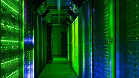 A brightly lit corridor in a data center features rows of server racks glowing with green and blue LED lights.
