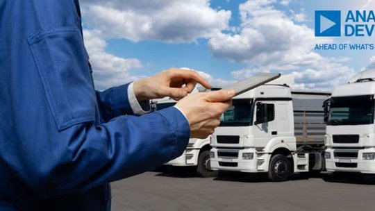 파란색 재킷을 입은 사람이 반쯤 흐린 하늘 아래 야외에 주차된 여러 대의 흰색 트럭 앞에서 태블릿 장치를 작동하고 있습니다. 오른쪽 상단에는 Analog Devices 로고와 'AHEAD OF WHAT'S POSSIBLE'이라는 문구가 명확하게 보입니다.