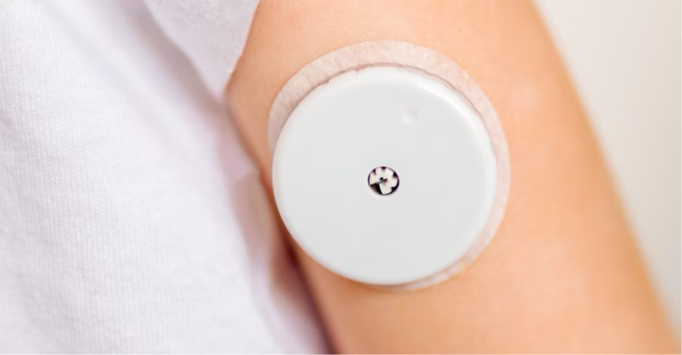 A circular wearable medical sensor is attached to the upper arm of a person wearing a white shirt. The device is secured with an adhesive patch and features a small central port.