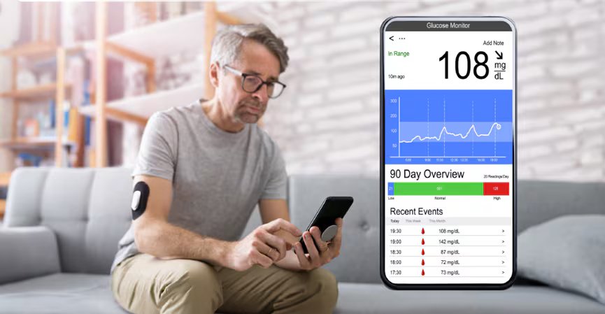 An adult man sits on a sofa in a modern living room, checking his smartphone. He wears a glucose monitoring sensor on his upper arm. A close-up of a mobile app interface displays glucose data, including a reading of 108 mg/dL and a 90-day overview chart.