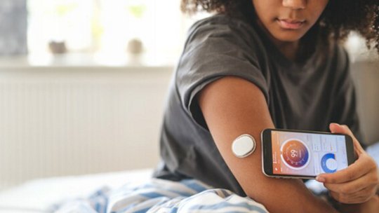 A young woman sits on a bed and uses her smartphone to scan a circular glucose monitor attached to her upper arm. The phone screen displays a colorful interface with a numeric glucose reading.