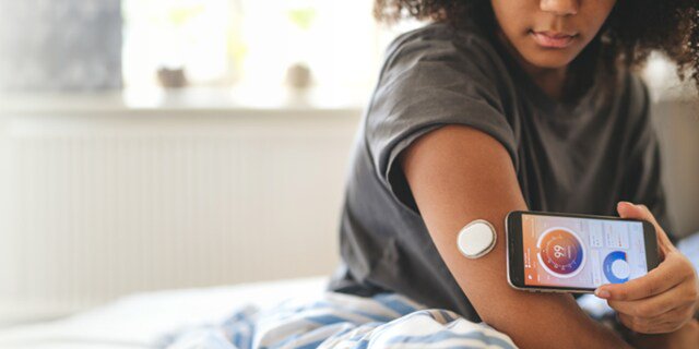 A young woman sits on a bed and uses her smartphone to scan a circular glucose monitor attached to her upper arm. The phone screen displays a colorful interface with a numeric glucose reading.