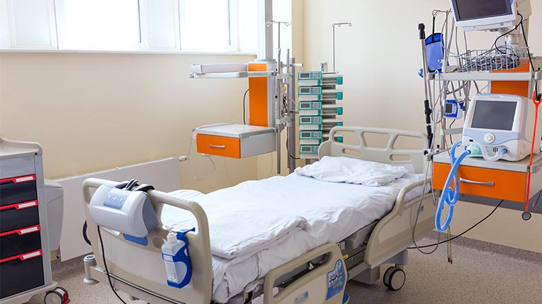 A hospital room features a single adjustable bed surrounded by advanced medical equipment and monitors. The setting is clean and well-lit, with neutral walls and organized storage units. Various devices are visible, including a ventilator and infusion pumps, emphasizing a clinical and sterile environment. No people are present, and no visible text or numbers can be seen on the equipment.