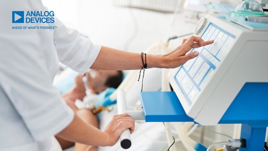의료 전문가가 터치스크린 인터페이스를 갖춘 최신 의료 모니터링 장치에서 설정을 조정하고 있습니다. 배경에는 신생아가 병원 침대에 누워 있어 신생아 치료 환경을 시사합니다. 장면에는 깨끗한 흰색 및 파란색의 임상 장비가 배치되어 첨단 의료 기술을 강조합니다. 왼쪽 상단에는 Analog Devices 마크와 “Ahead of What’s Possible” 문구가 표시되어 있습니다.