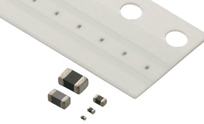 The image shows several tiny surface mount electronic components arranged in front of a white carrier tape. The components vary in size, with the largest in the foreground and progressively smaller parts behind it. The carrier tape features evenly spaced pockets and two circular indexing holes along one edge. The scene is clean and minimal, emphasizing precision manufacturing and miniaturized electronics.