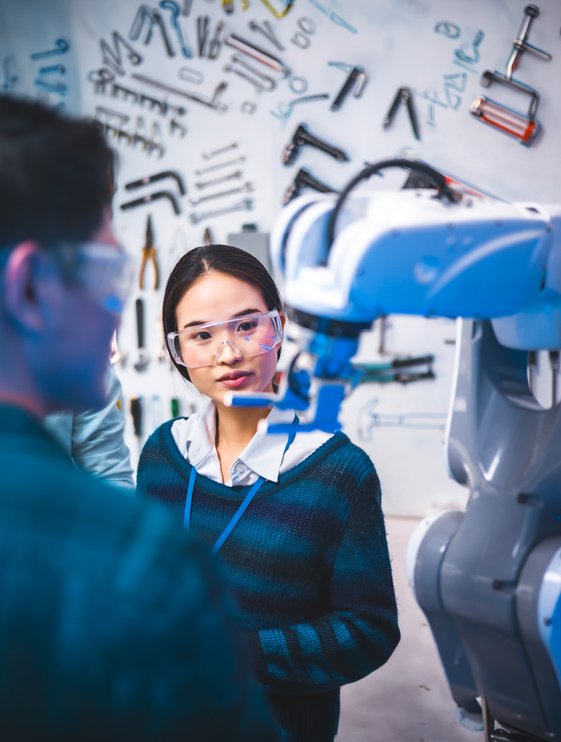 A young engineer wearing safety glasses observes an industrial robotic arm in a workshop or lab. Another person stands in the foreground, partially visible, engaged in the discussion. The background wall is covered with various tools, emphasizing a technical and mechanical environment. Cool lighting and sharp focus highlight the advanced machinery and collaborative engineering work.