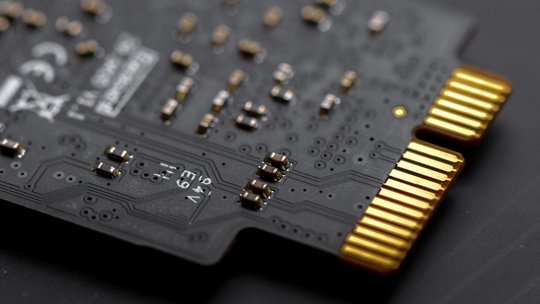 Una vista dettagliata di una scheda elettronica nera con connettori placcati in oro. La scheda presenta componenti complessi e dettagli di saldatura, mettendo in risalto l'ingegneria di precisione. Il testo visibile include '12V' e altre marcature tecniche, evidenziando le sue specifiche elettriche.