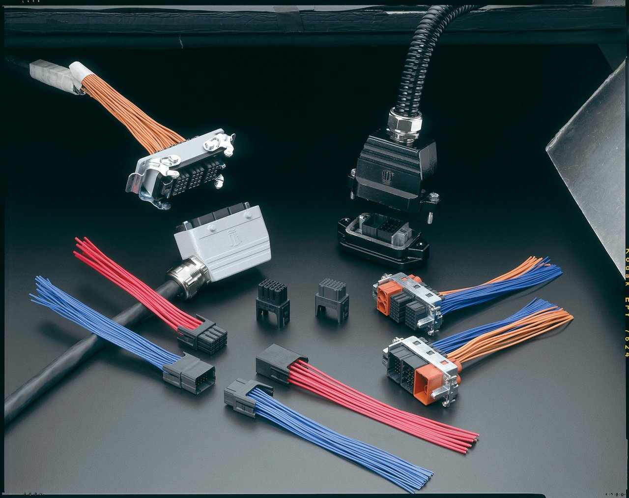 The image shows a collection of industrial electrical connectors arranged on a dark surface. Multiple plug and socket components are attached to bundles of colored wires, including orange, blue, and red leads. Some connectors are mounted in metal housings, while others are standalone plastic blocks. The scene highlights the variety of connector types and cable assemblies used in electrical or electronic installations.