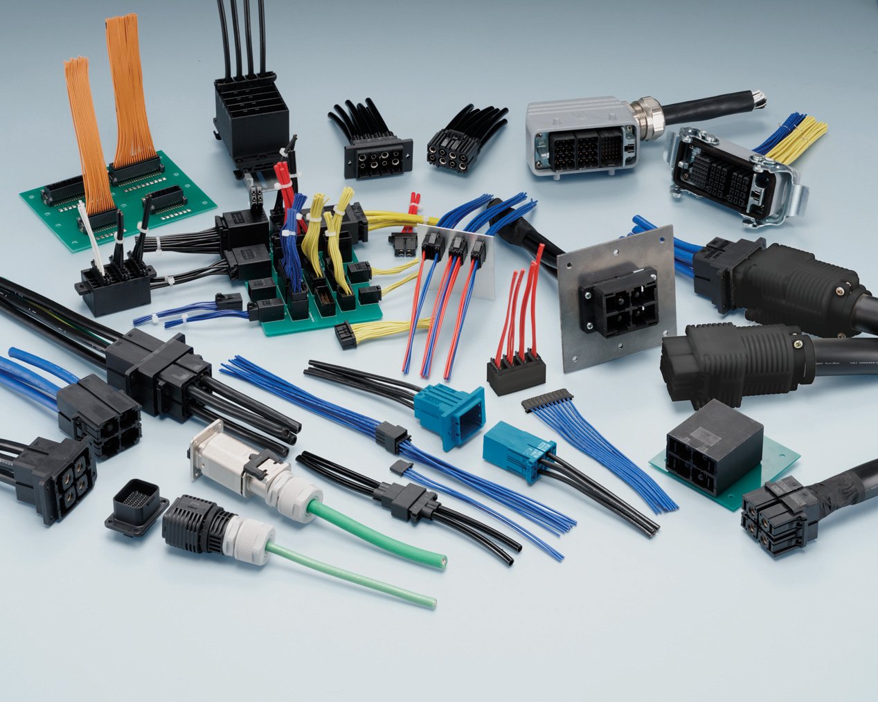 A variety of industrial electrical connectors and cables are arranged on a light surface. The components include plug housings, sockets, and terminal blocks in different shapes and sizes. Color-coded wires in blue, red, yellow, green, and orange are attached to several connectors and circuit boards. The image highlights the diversity of connection types used in electrical and electronic installations.