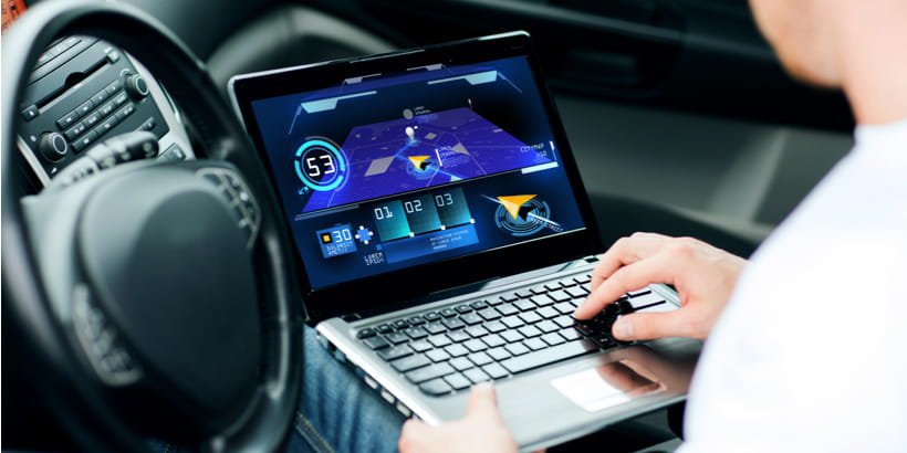 A person is seated inside a car, working on a laptop that displays a digital dashboard interface. 