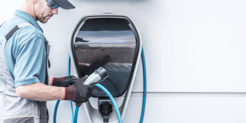 A technician in work attire is connecting a charging cable to an electric vehicle charging station. 