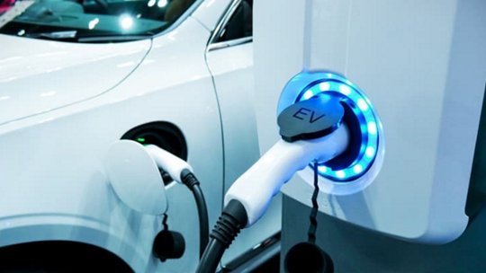A white electric vehicle is connected to a charging station, showcasing a glowing blue EV plug