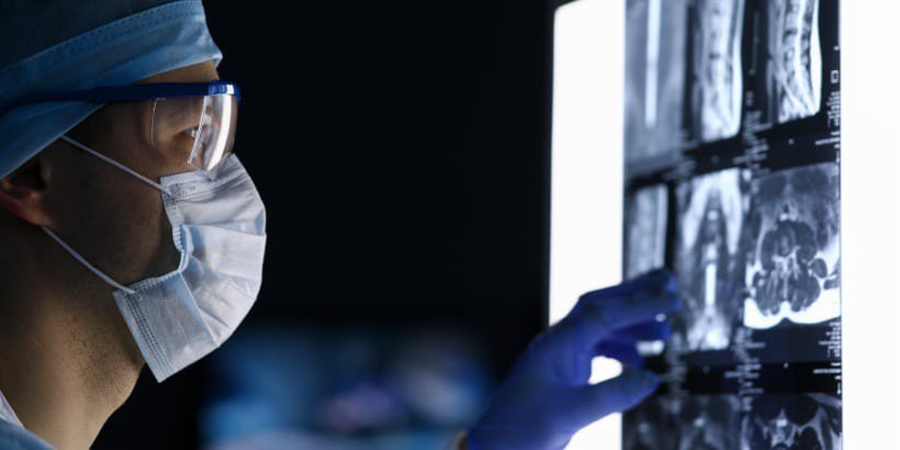 A healthcare professional wearing surgical attire examines X-ray images on a backlit screen in a clinical setting.