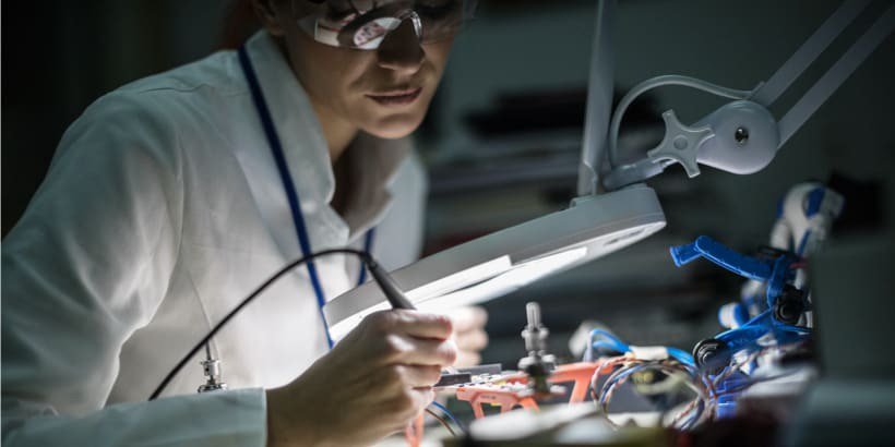 A technician in a white lab coat is working on electronic components under a magnifying lamp..