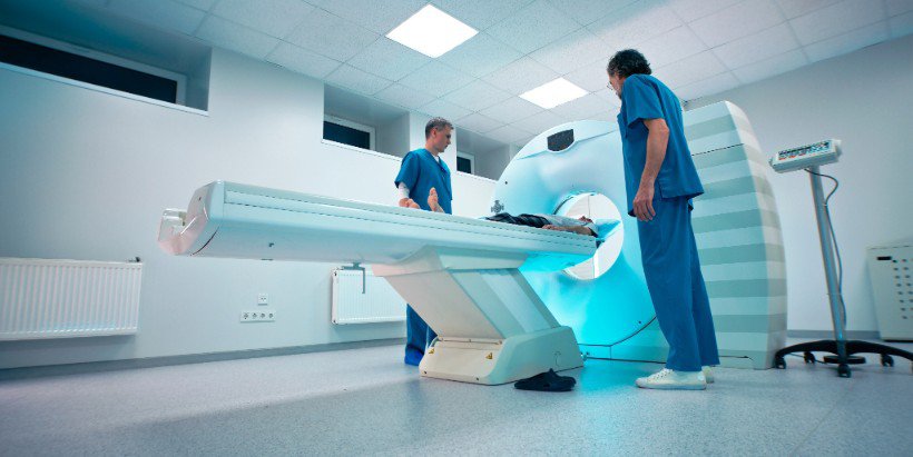 Two healthcare professionals in blue scrubs operate a modern medical imaging machine in a clinical setting.