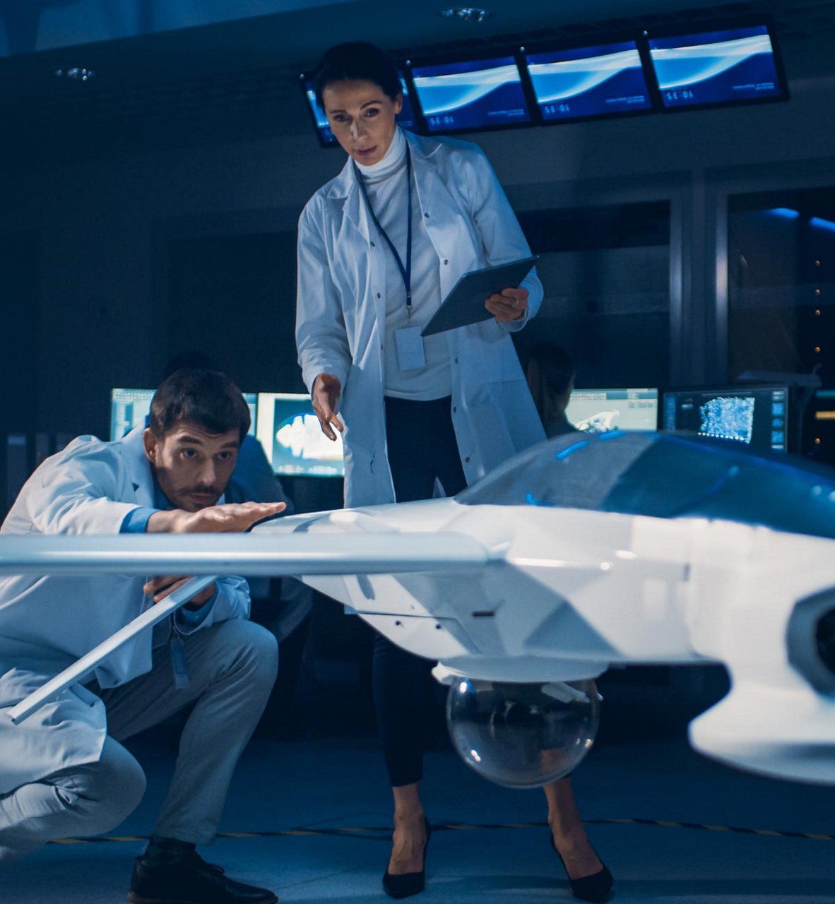 Dos ingenieros con batas de laboratorio examinan un gran dron aéreo no tripulado en una sala de control de alta tecnología.