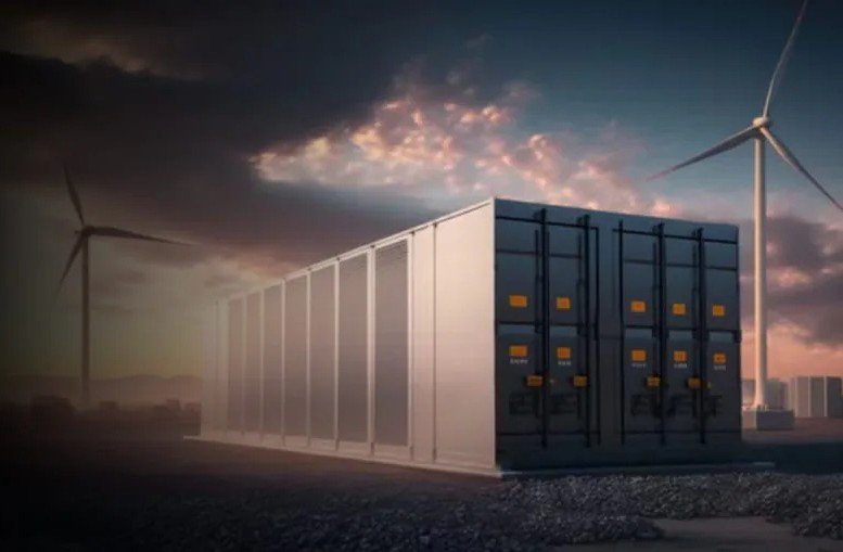 A large industrial battery storage unit is positioned outdoors, with wind turbines visible in the background under a dramatic sunset sky.