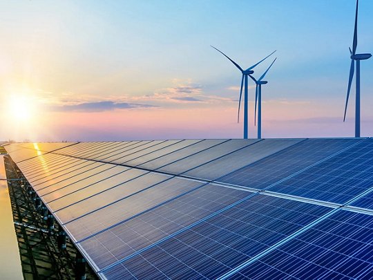A large array of solar panels is prominently displayed in the foreground, reflecting the early morning sunlight. In the background, several wind turbines stand tall against a clear sky, highlighting a renewable energy landscape.
