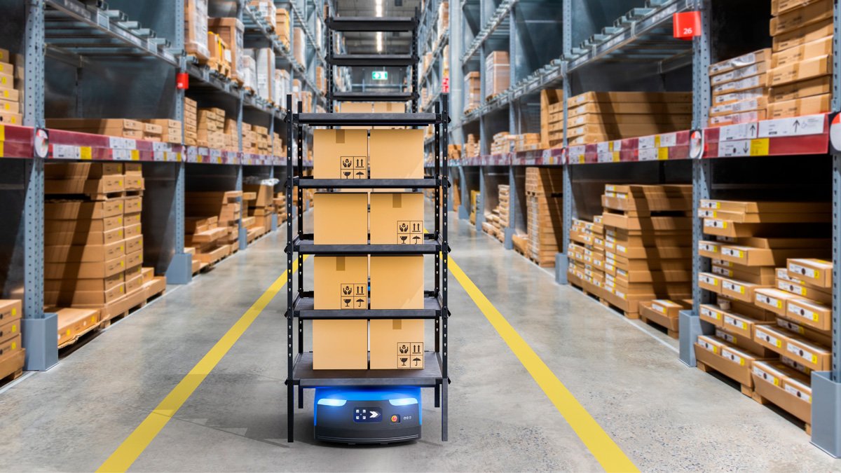 An autonomous mobile robot carries multiple shelves stacked with cardboard boxes down an aisle in a large warehouse. 