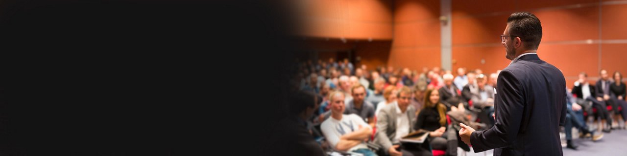 A man in a suit stands in front of a seated audience, delivering a presentation in a modern conference hall.