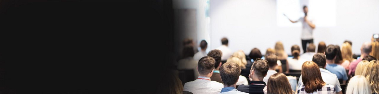 A presenter stands at the front of a room addressing a seated audience. 