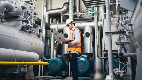 Ingénieur industriel en gilet de sécurité utilisant un ordinateur portable près d'une grande machine grise et métallique.