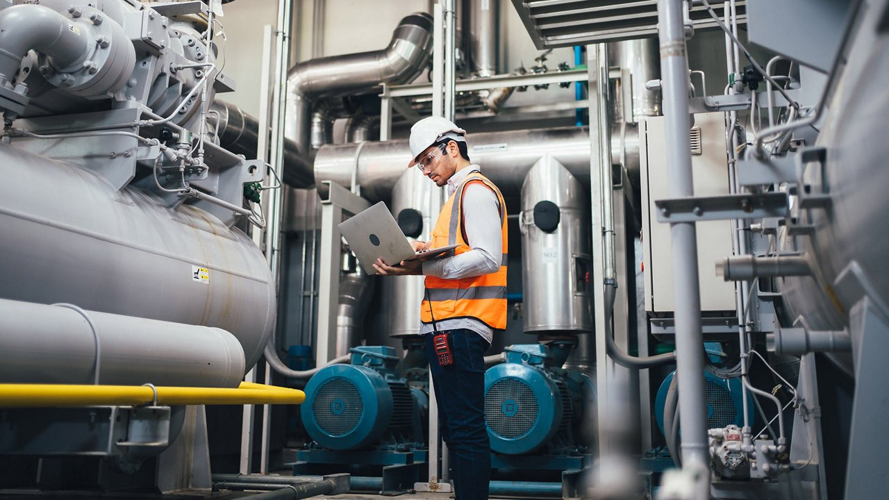Ingénieur industriel en gilet de sécurité utilisant un ordinateur portable près d'une grande machinerie grise et métallique.