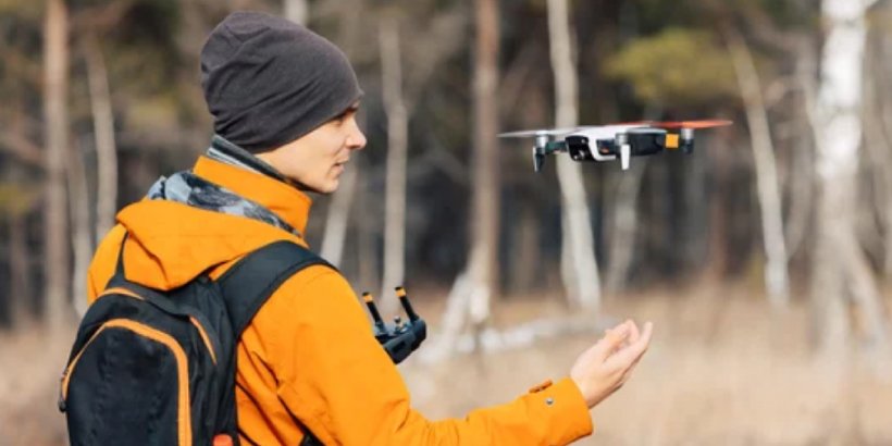 A person wearing an orange jacket and backpack is seen controlling a drone in a natural outdoor environment. 