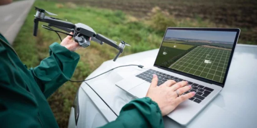 A person operates a drone while using a laptop on the hood of a car in a rural field.