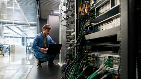 현대식 서버실에서 한 기술자가 네트워크 장비 랙 사이에 노트북으로 작업하며 웅크리고 있다.