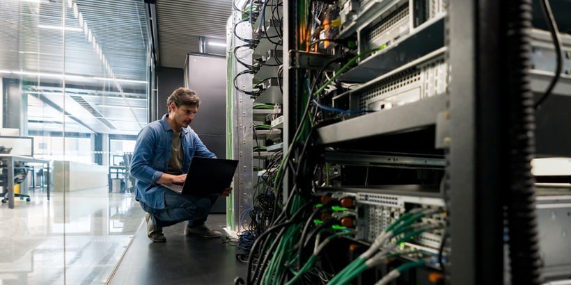 한 기술자가 현대적인 서버 룸에서 네트워크 장비 랙 사이에 노트북으로 작업하며 쪼그리고 앉아 있습니다.