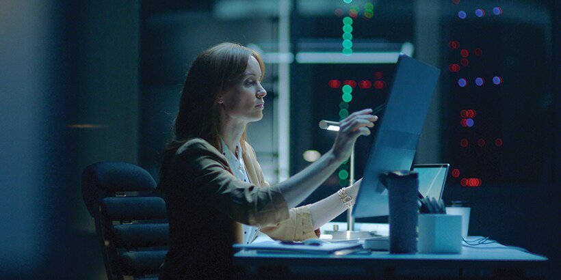 A woman is seated at a desk in a dimly lit server room, reviewing documents under focused lighting