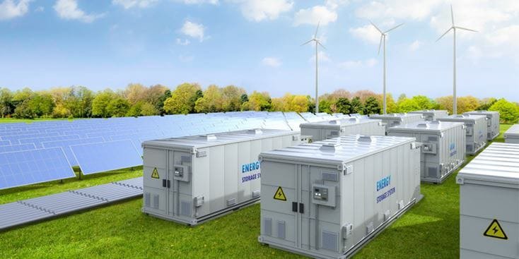 A row of energy storage containers is positioned in a green field surrounded by solar panels and wind turbines