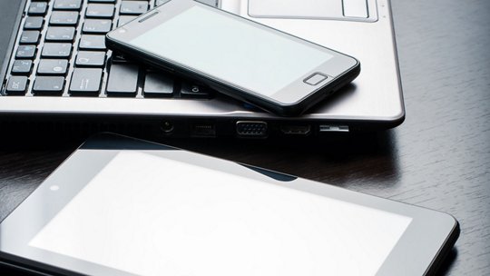A close-up of a laptop, smartphone, and tablet placed on a dark wooden desk. 