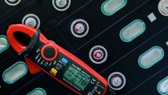 Un medidor digital de pinza rojo y negro está colocado sobre una cuadrícula de celdas de batería cilíndricas.