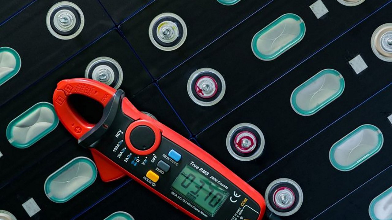Un ampèremètre numérique rouge est placé sur une grille de cellules de batterie, mettant en valeur son affichage de mesure indiquant '03.710'. L'environnement semble être technique ou industriel, axé sur l'électronique ou le stockage d'énergie.
