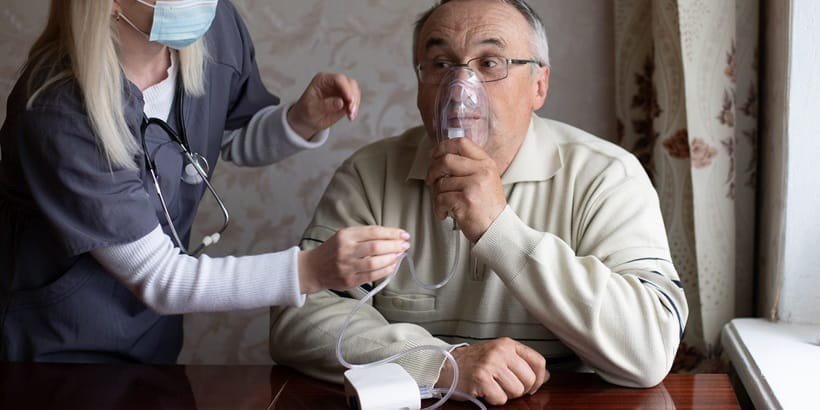 A healthcare professional wearing a mask is assisting a patient with a medical device in a home setting