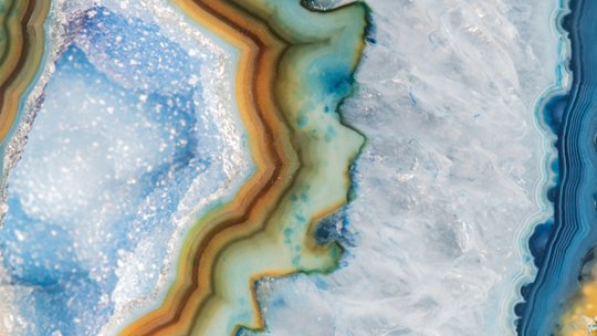 A detailed macro shot showcasing the intricate layers of a polished agate stone. The image highlights vibrant hues of blue, green, and amber with crystalline textures. 