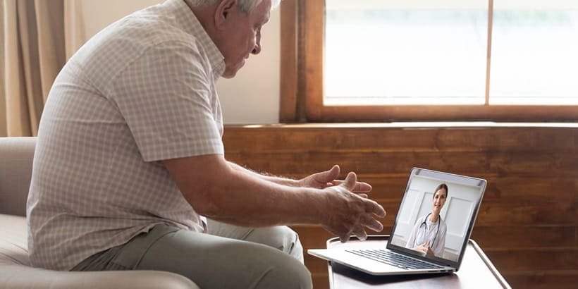 An elderly man sits in a cozy living room setting, engaging in a virtual consultation with a doctor displayed on a laptop screen