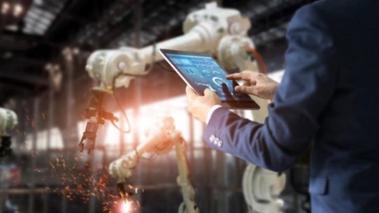 A professional in a suit operates a tablet device in a modern industrial setting with robotic arms. 