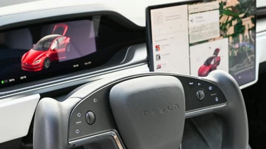 테슬라 자동차 내부를 클로즈업으로 보여주며 미래지향적인 스티어링 휠과 듀얼 디지털 디스플레이를 강조합니다.