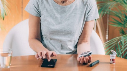 A middle-aged woman sits at a wooden table, using a glucose monitoring device and a smartphone. 