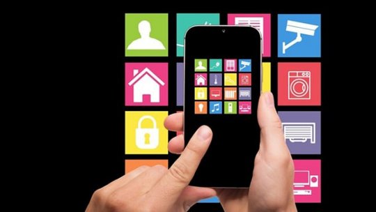 A close-up of hands holding a smartphone displaying colorful home automation icons on the screen. 