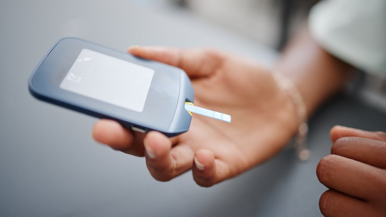 A person holds a handheld blood glucose meter with a test strip inserted at the bottom.